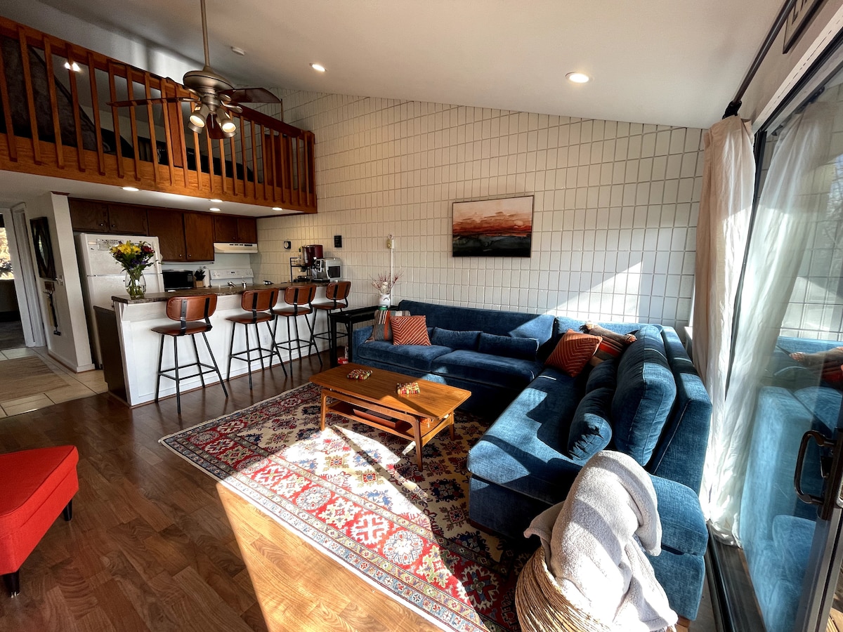 An open living area is highlighted by a large blue sectional sofa, a wooden coffee table, and a vibrant area rug. Natural light fills the space through expansive glass doors. A bar with four stools is visible in the background, leading to a well-equipped kitchen.