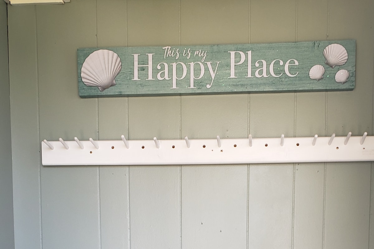 A sign reading 'This is my Happy Place' hangs above a row of white hooks on a textured green wall, adorned with seashell graphics. The scene evokes a coastal charm, inviting guests to feel at home in this coastal cottage.