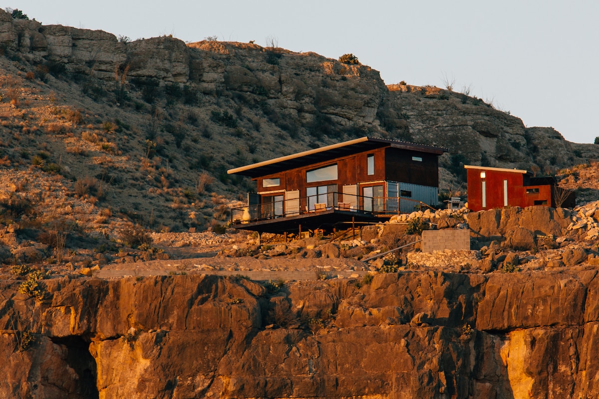 A modern cabin-style home perches on the edge of a limestone cliff, showcasing a blend of wood and metal exteriors. Large windows and a cantilevered deck offer expansive views of the surrounding desert landscape and geological formations.
