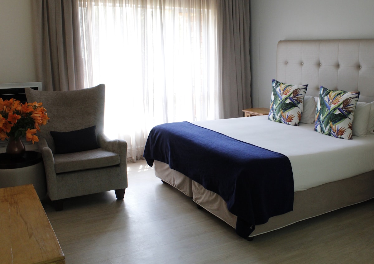 A spacious room features a large bed with a dark blue blanket and decorative throw pillows showcasing tropical patterns. A comfortable armchair is positioned near the window, which is dressed in light, flowing curtains, allowing natural light to brighten the space.