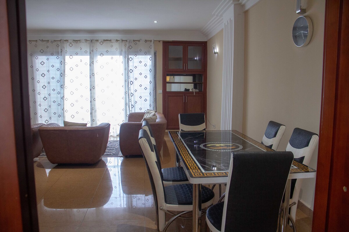 An open living area is showcased, featuring a dining table surrounded by six chairs. A comfortable seating arrangement with two armchairs and a sofa is visible. Large windows allow natural light to fill the space, complemented by light-colored curtains and a decorative wall clock.
