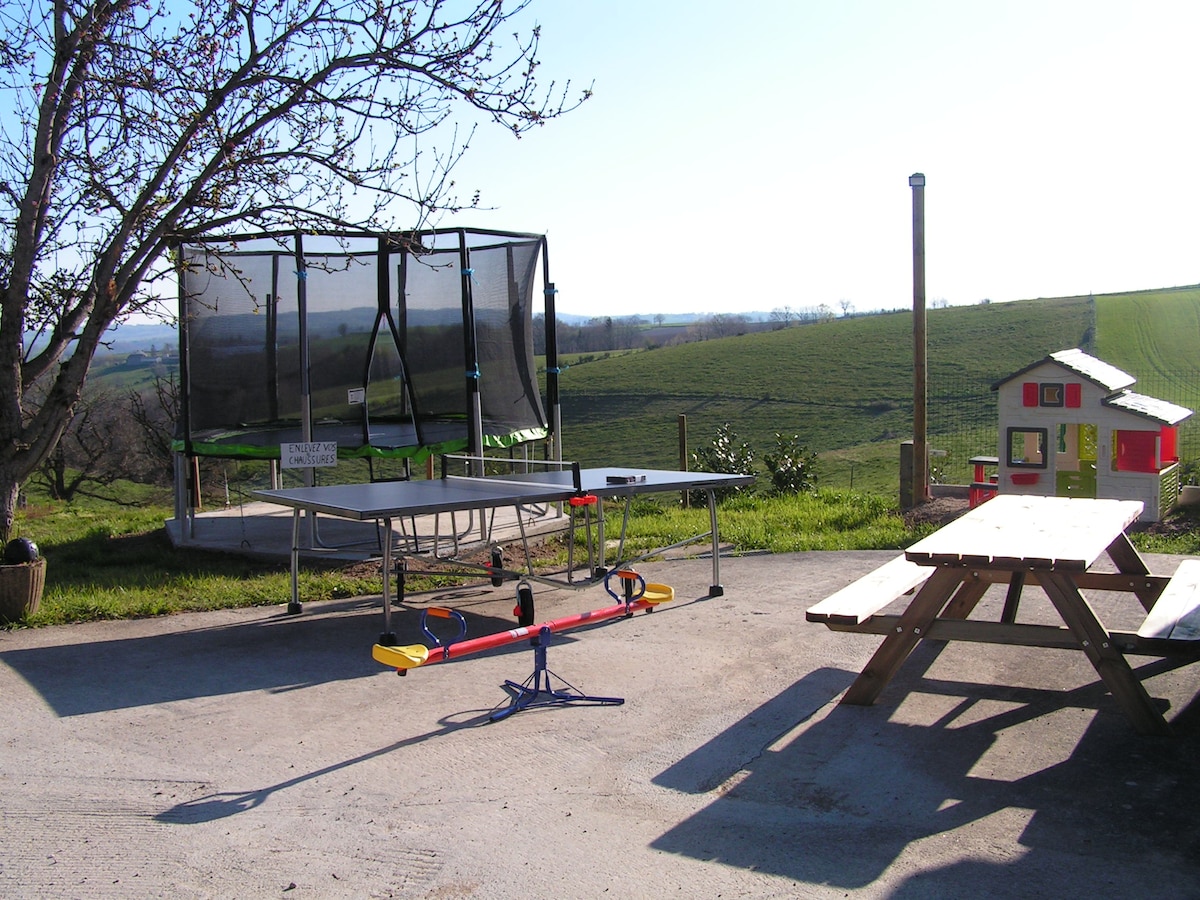 An outdoor space features a trampoline, a balancing seesaw, and a playhouse, surrounded by greenery. A picnic table provides a gathering area, while a tree casts light shade. The open landscape stretches in the background, offering a view of rolling hills.