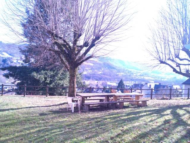 A spacious outdoor area features a wooden picnic table under a large tree, surrounded by a grassy lawn. The scene is accented by a panoramic view of rolling hills in the distance and is bordered by a wooden fence, providing a peaceful environment.