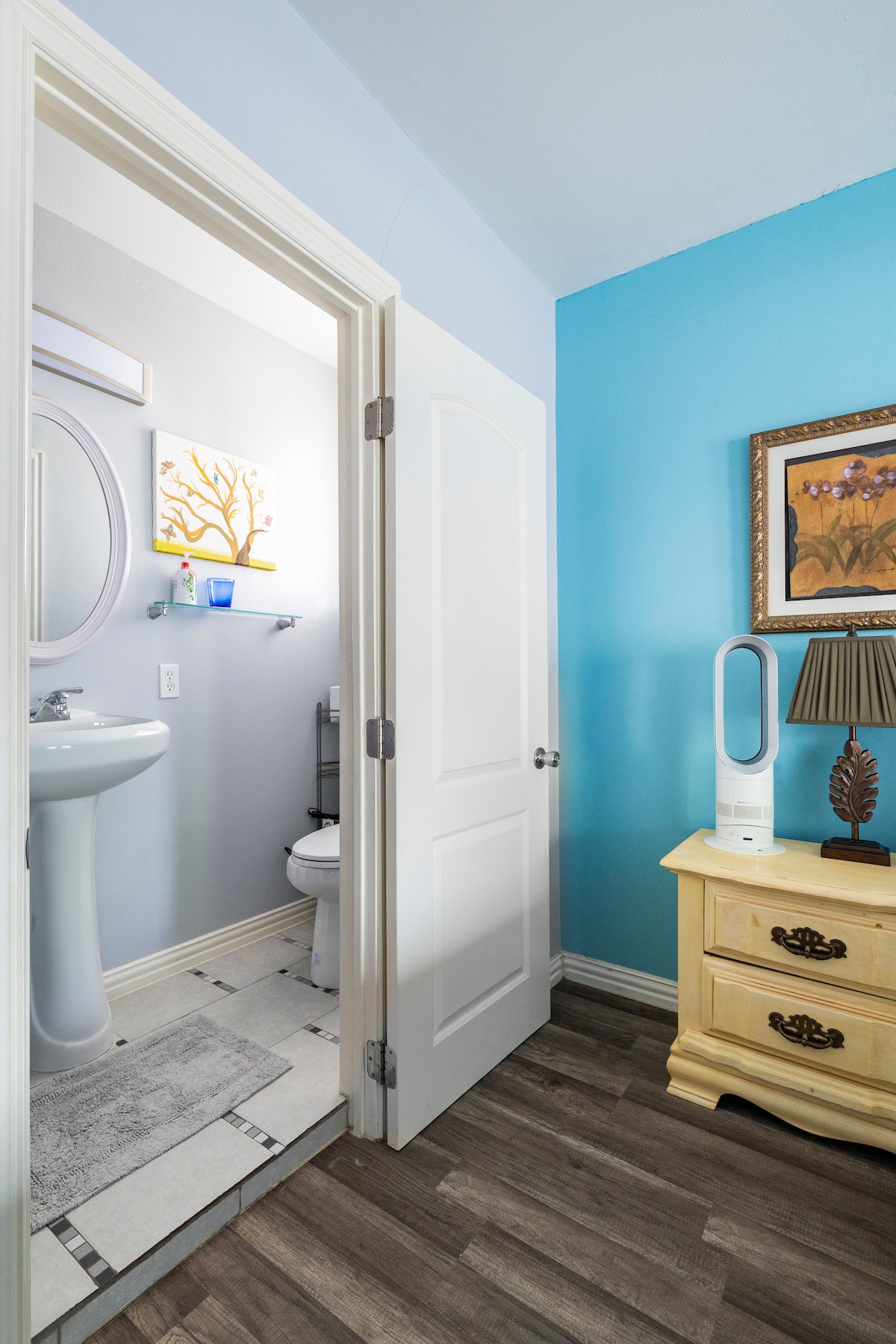 A brightly painted bathroom features a white pedestal sink and a toilet. Soft blue walls enhance the space, while a decorative piece hangs nearby. A small wooden nightstand with a lamp is positioned outside the bathroom door, contributing to a cozy atmosphere.