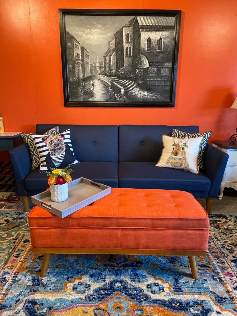 A cozy seating area features a navy blue couch adorned with decorative pillows. An orange tufted ottoman sits at the center, holding a tray with fruit. A vibrant area rug adds warmth, complemented by an art piece depicting a cityscape on the wall.
