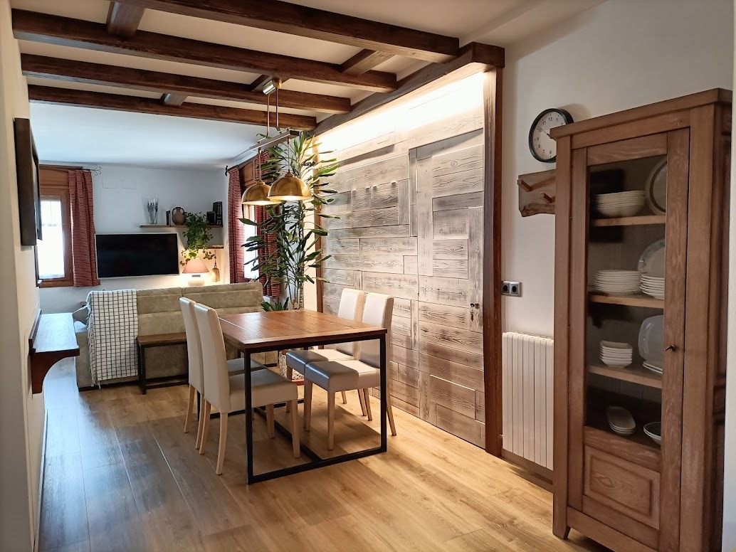 The dining area features a wooden table surrounded by four light-colored chairs. A comfortable sofa is positioned near a flat-screen TV on the wall. Natural light fills the space, highlighting the wooden beams of the ceiling and decorative plants along the walls.