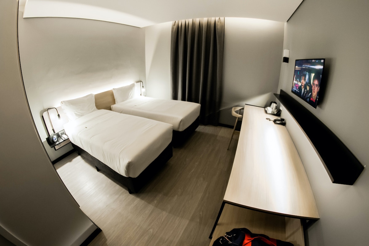 A modern guestroom features two spacious single beds, each with minimalistic white bedding. A large flat-screen TV is mounted on the wall, while a wooden desk and chair sit nearby. Soft lighting is provided by wall lamps, and dark curtains frame the window.