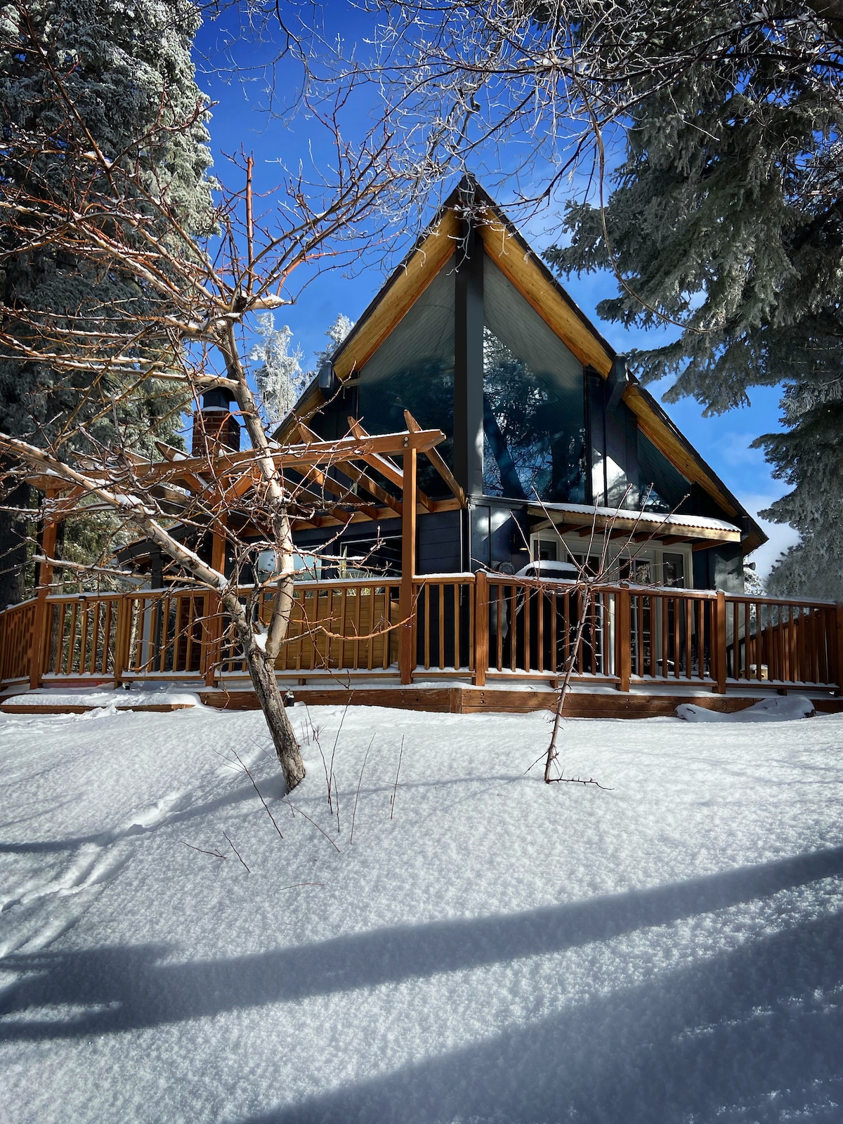 The charming A-frame cabin is surrounded by a snowy landscape, showcasing a spacious wooden deck and tall glass windows that reflect natural light. Towering trees and soft snow cover the ground, creating a serene mountain setting.