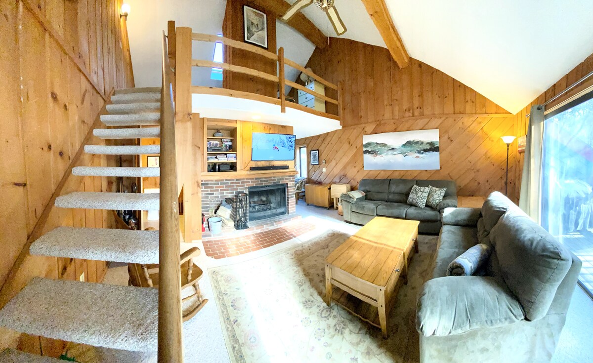 The spacious living area features natural wood paneling and high vaulted ceilings. A comfortable seating arrangement includes a sectional sofa and a coffee table, located near a brick fireplace. A staircase leads to a loft area, while a large window offers views of the outdoors.
