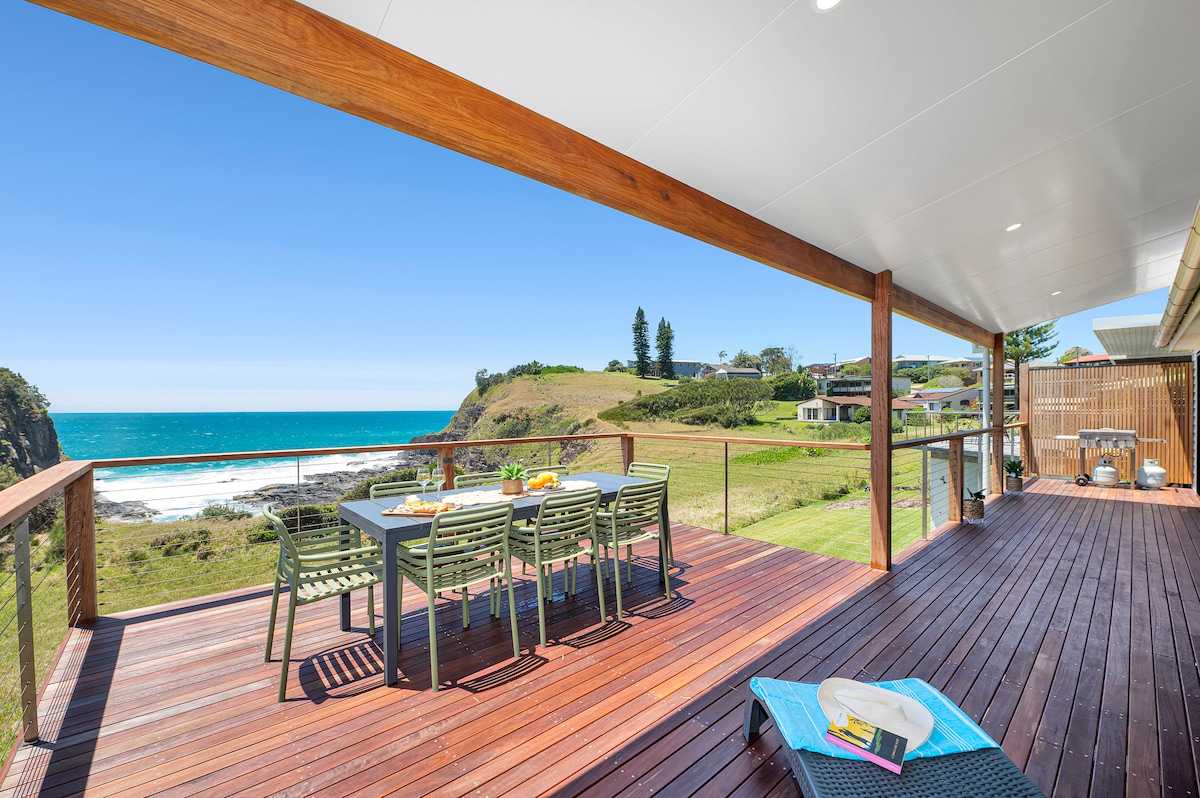 A spacious deck features a generous dining table surrounded by modern chairs, overlooking a vibrant ocean view. The shoreline is visible in the distance, complemented by clear blue skies. Sun loungers are positioned nearby, inviting relaxation in the coastal atmosphere.