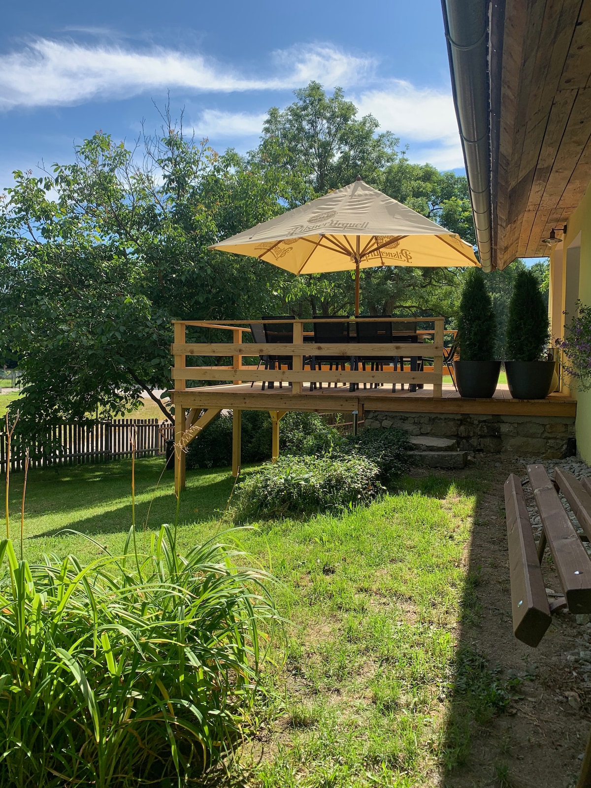 An elevated wooden deck features a dining area, shaded by a large umbrella. Green grass surrounds the deck, complemented by tall shrubs and trees providing a natural setting. A bench is positioned nearby, enhancing the outdoor space.