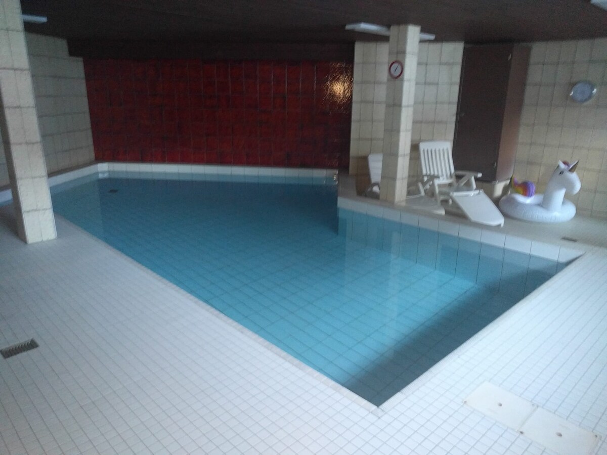 An indoor swimming pool is visible, featuring a clear blue water surface that reflects light. White tiled walls and floor surround the pool area, while a red accent wall adds contrast. A few lounge chairs are positioned beside the pool, enhancing the relaxation space.