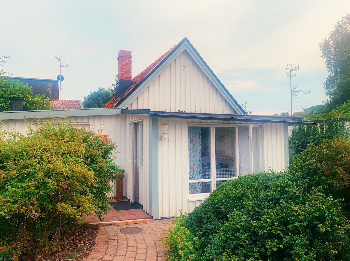 The exterior of a quaint white cottage is framed by lush greenery and a curved pathway. A chimney and a gabled roof add character to the building, while large windows allow glimpses of the inviting interior. Soft daylight creates a welcoming appearance.