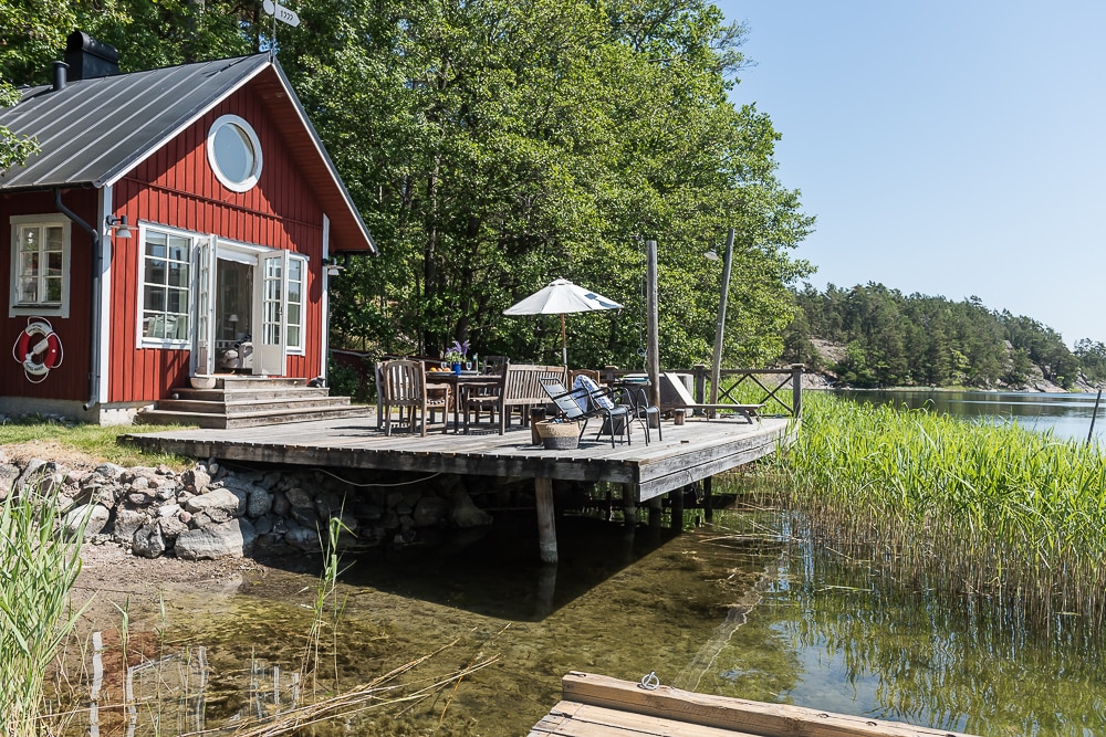 A red wooden cabin is positioned next to a dock, surrounded by lush green trees and water. Outdoor furniture, including a table and chairs, is set up on the spacious deck, inviting relaxation. The serene environment is complemented by the clear water and natural vegetation.