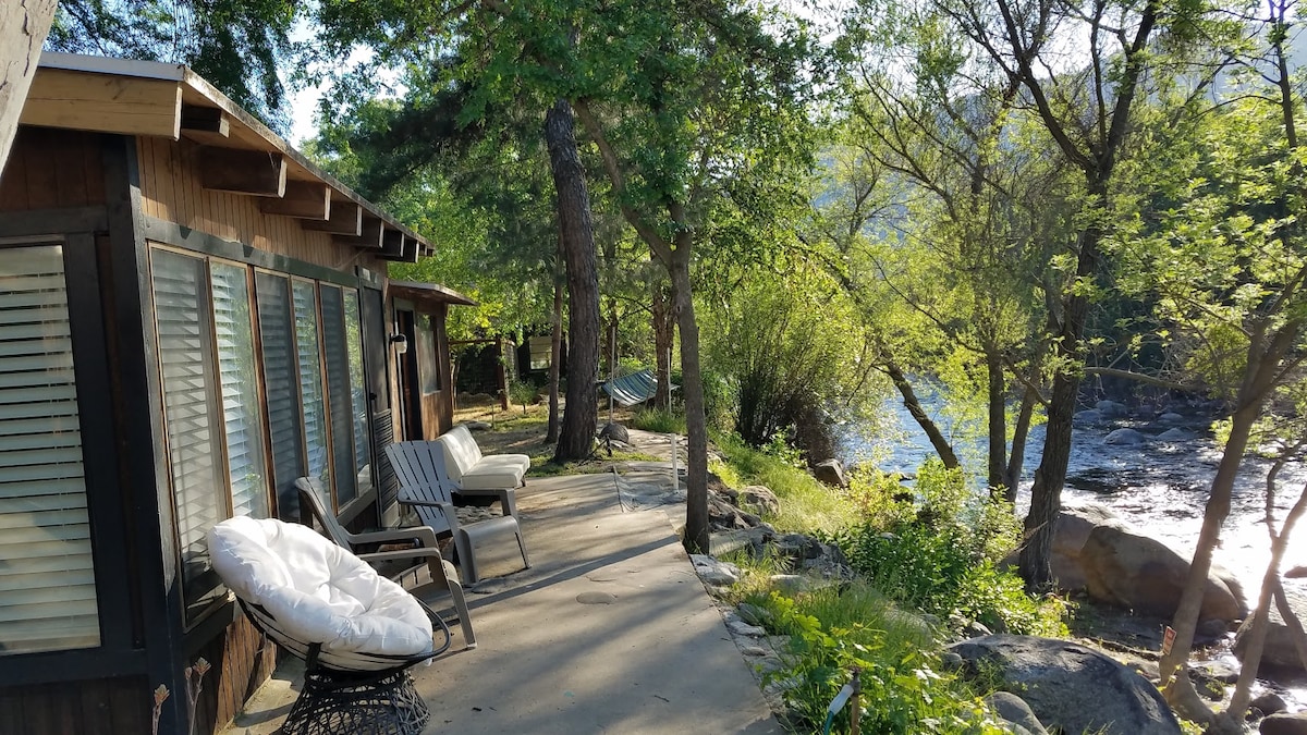 A concrete pathway runs alongside the river, flanked by trees and greenery. Comfortable seating options, including a lounge chair and cushioned chair, are positioned to enjoy the view. Sunlight filters through the leaves, casting dappled shadows on the ground.