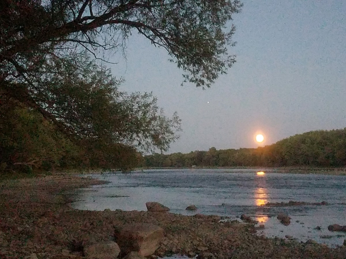 A calm river scene captures a full moon rising above the water, casting a soft glow. The shoreline is lined with large rocks, and trees provide a natural frame to the tranquil setting. Reflections shimmer on the surface, enhancing the serene atmosphere.