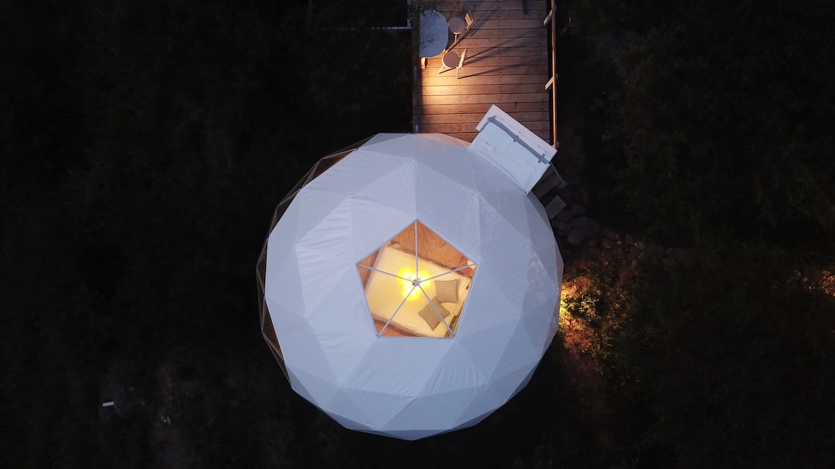 An aerial view of a geodesic dome structure is displayed, set amongst greenery. A wooden deck leads to the dome, which features a skylight at the top for natural light. Soft illumination is visible inside, providing a warm glow in the surroundings.