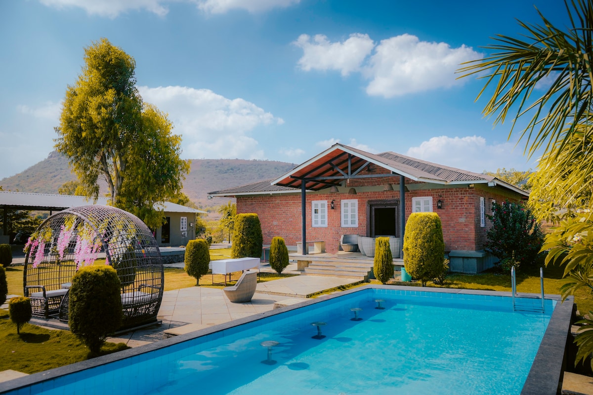 A spacious villa is surrounded by well-maintained lawns, notably featuring a vibrant blue pool in the foreground. Comfortable lounge areas are positioned nearby, with stylish seating and greenery enhancing the setting. A distinctive round structure stands to the left, adding an element of interest to the landscape.