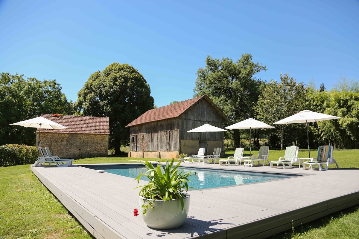 A sunlit saltwater swimming pool is surrounded by spacious decking and elegant loungers. Umbrellas provide shade, while a charming wooden barn is visible in the background, nestled among lush green trees and open countryside.