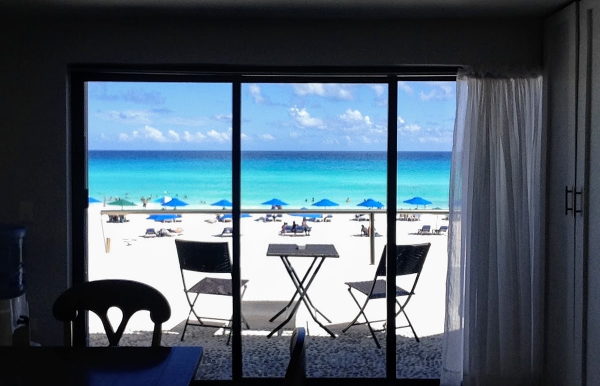 A clear view of the Caribbean Sea is framed by large glass doors. Two chairs and a small table are set against the backdrop of pristine sandy shore and umbrellas, defining a serene outdoor space that invites relaxation.