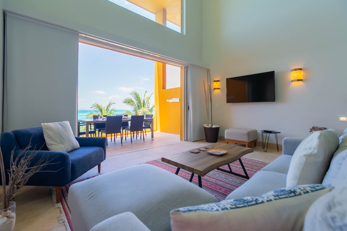 A spacious living area is showcased, featuring comfortable seating and a wooden coffee table. Large sliding doors open to a bright outdoor dining space adorned with palm trees, inviting views of turquoise waters, and a vibrant yellow wall that adds warmth.