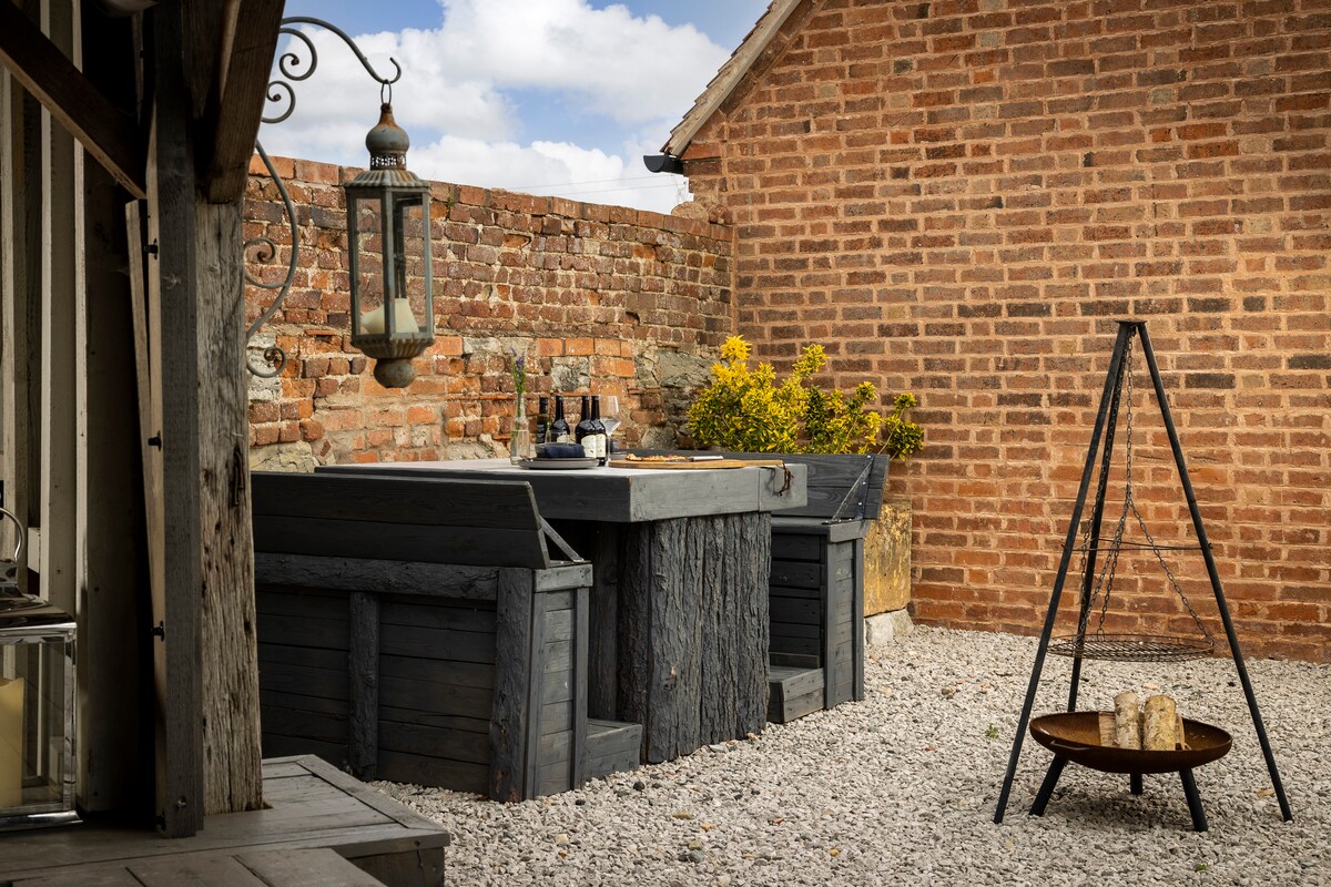 A wood-fired hot tub is surrounded by a stone and gravel area, nestled against a rustic brick wall. A small table is set nearby with two glasses and a bottle. A lantern hangs from the structure, providing soft ambient light.