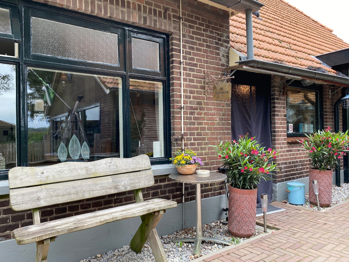 An inviting outdoor area features a wooden bench next to a small table adorned with colorful flowers. Flower pots with vibrant greenery add charm, while textured brick walls and large windows provide a sense of connection to the scenery beyond.