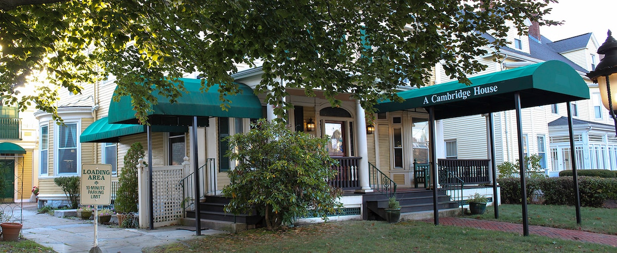 A charming Victorian house is framed by lush trees, featuring green awnings over the entrance. The porch is accessible, and a welcoming sign indicates the property's name. A pathway leads from the street to the front door, enhancing the inviting exterior.