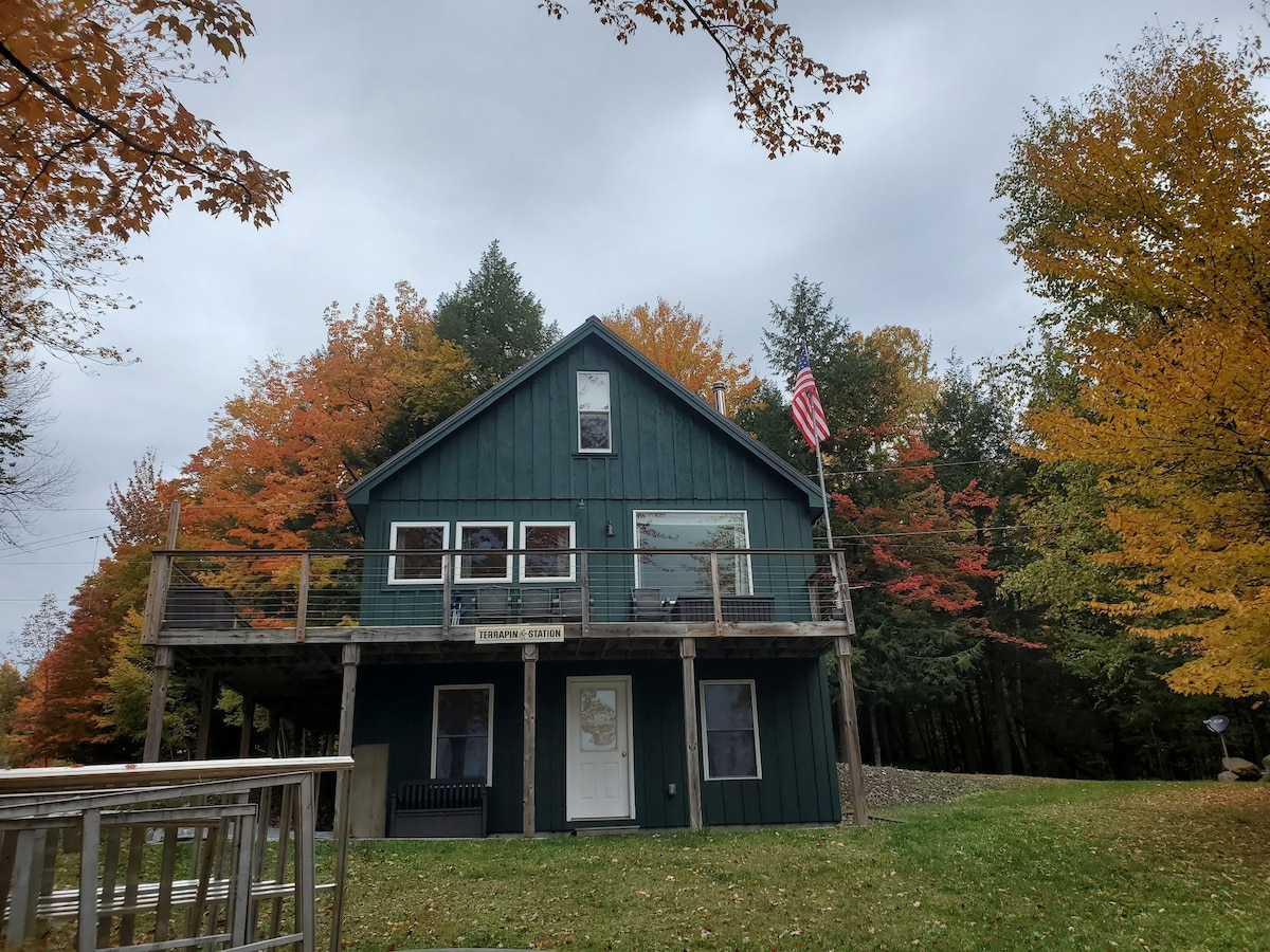 The cabin presents a two-story structure with a green exterior and multiple large windows, providing natural light. A wooden deck extends from the front, adorned with a railing. Surrounding foliage displays vibrant autumn colors, while an American flag is gently displayed on the front.