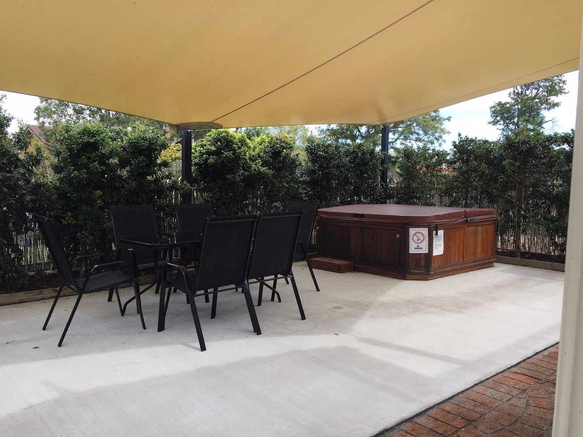 An outdoor space is sheltered by a large, beige awning. A wooden hot tub is positioned against a backdrop of greenery. Black chairs are arranged around a table on a paved surface, providing a designated area for relaxation and socializing.