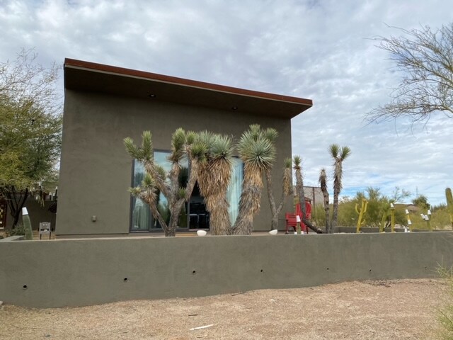 A modern house exterior features large windows framed by desert landscaping, including various palm and cacti species. The earthy toned wall contrasts with the red accent chairs positioned on the porch, creating a harmonious connection with the surrounding arid environment.