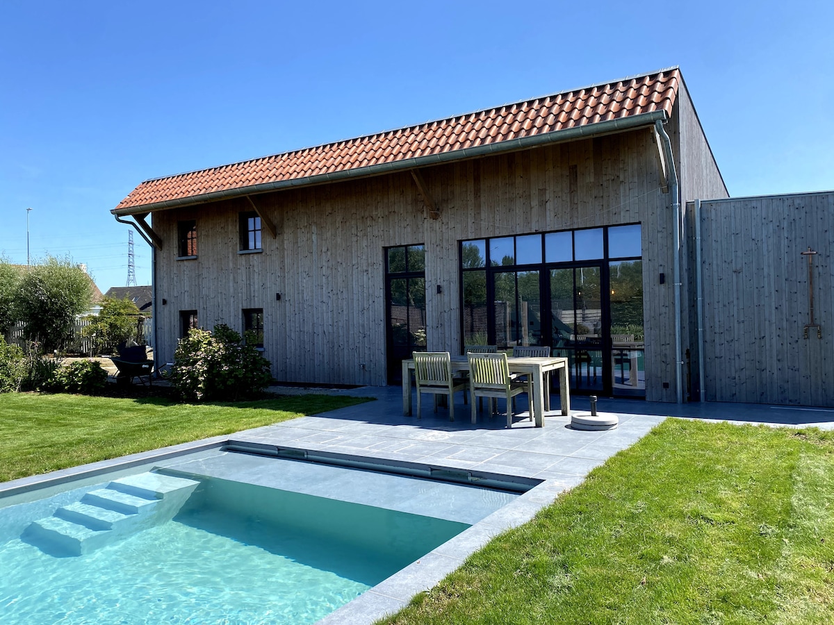 A modern wooden structure features large glass windows, allowing natural light to fill the interior. A patio with outdoor seating is situated next to a well-maintained lawn and a heated pool, creating an inviting space for relaxation and outdoor activities.