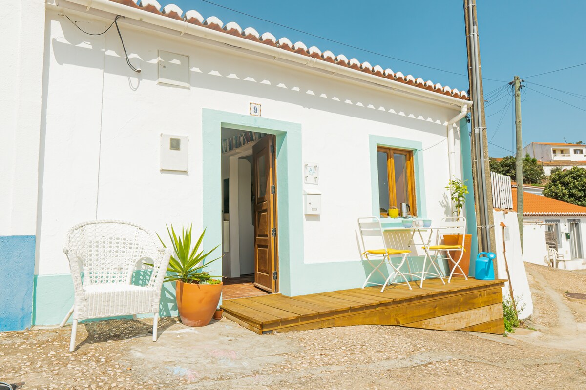 The exterior of the house is captured in bright sunlight, featuring a welcoming entrance with a wooden deck. Two windows with wooden frames allow natural light to enter. A small table and two chairs sit outside, accompanied by a potted plant, enhancing the peaceful atmosphere.
