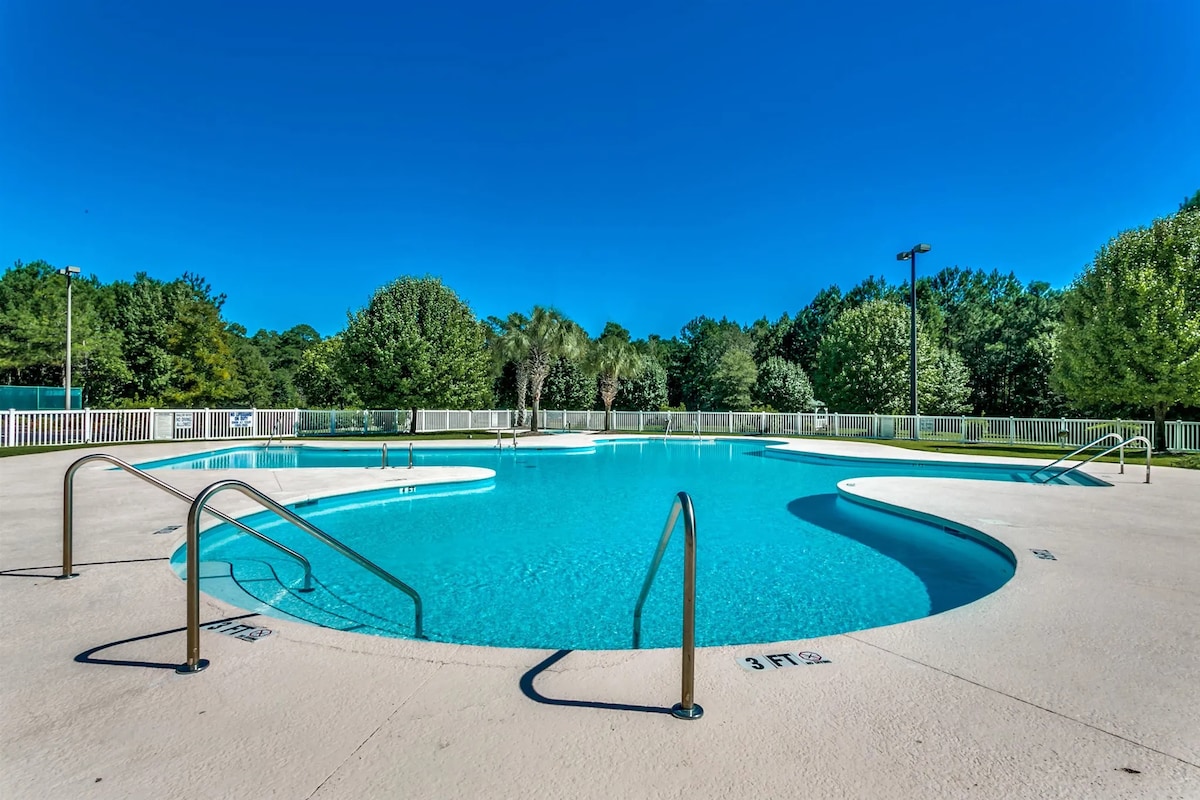 A large swimming pool is surrounded by a spacious deck and lush greenery. The water is clear and inviting, with gradual steps leading in. Sun loungers may be positioned nearby, offering a relaxing area to enjoy the outdoors.
