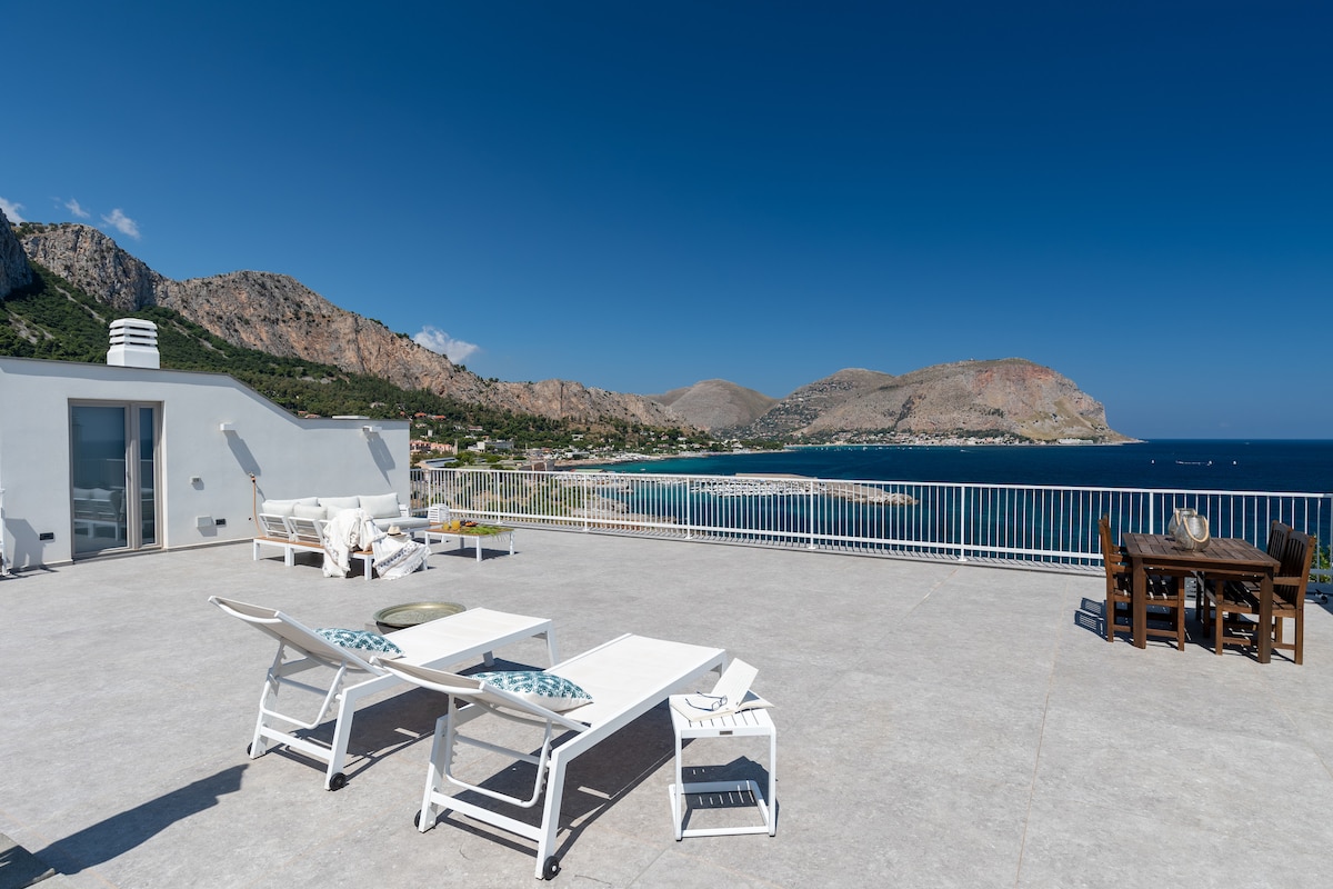 A spacious terrace features comfortable lounge chairs and a dining table, overlooking a breathtaking view of the coastline and mountains. The sun-drenched area offers ample space for relaxation and outdoor dining, complemented by clear blue skies and the glistening sea.