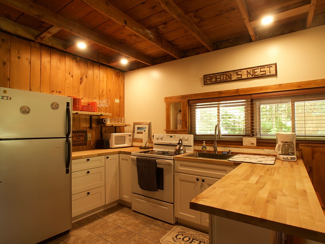 The kitchen features wooden cabinetry and natural wood beams along the ceiling. Silver appliances include a refrigerator and oven. A countertop offers ample preparation space, complemented by a window with horizontal blinds allowing light to enter. Simple kitchenware is neatly arranged, enhancing functionality.