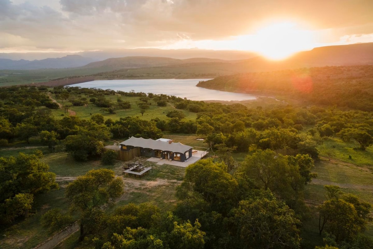 An aerial view captures a tranquil retreat surrounded by lush greenery, set against a backdrop of rolling hills and a serene lake. The golden hues of a sunset illuminate the area, highlighting the lodge with a spacious outdoor deck.
