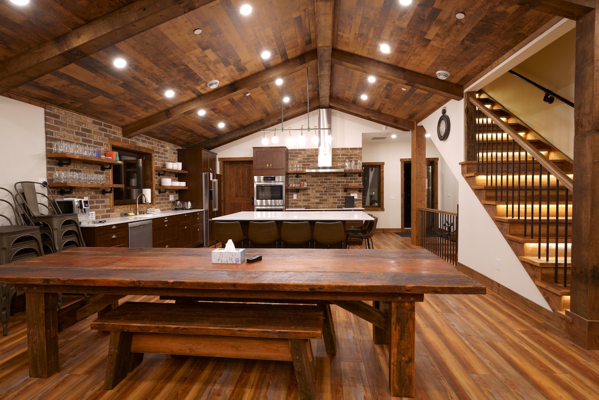 An expansive dining area features a large rustic wooden table surrounded by benches. The open kitchen is equipped with modern appliances, and warm wood accents create an inviting atmosphere. Skylights provide ample natural light, highlighting the unique ceiling design and stone accents.