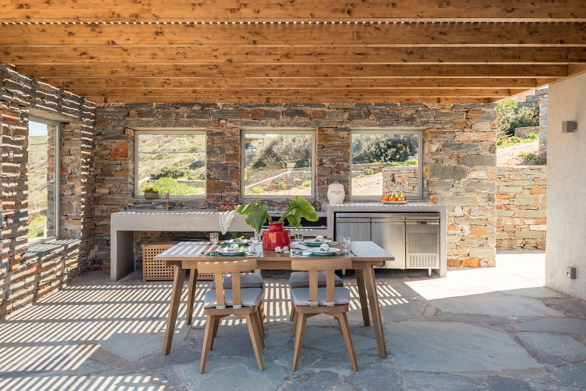An outdoor dining area features a wooden table set for a meal, surrounded by stone walls and large windows that provide a view of the landscape. A modern kitchen space is visible in the background, enhancing the functionality of the area.