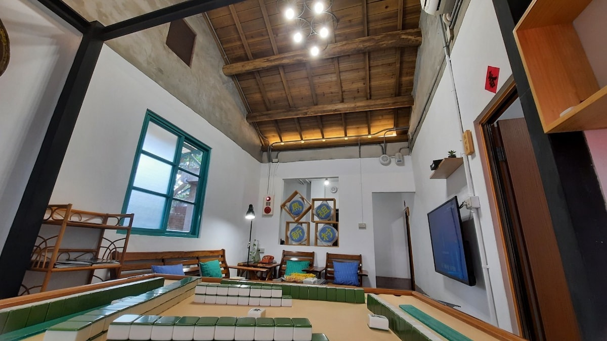 A spacious common area features high ceilings with exposed wooden beams, enhancing the sense of openness. Natural light filters in through large windows, illuminating a wooden seating area and a decorative screen. A modern television is mounted on the wall, providing entertainment options.