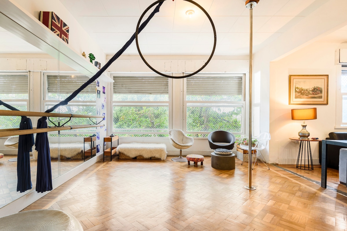 An expansive room designed for movement features a mirrored wall and various exercise apparatus, including a hoop and pole. Large windows allow natural light to fill the space, highlighting the warm wood flooring. Comfortable seating areas are arranged thoughtfully around the room.