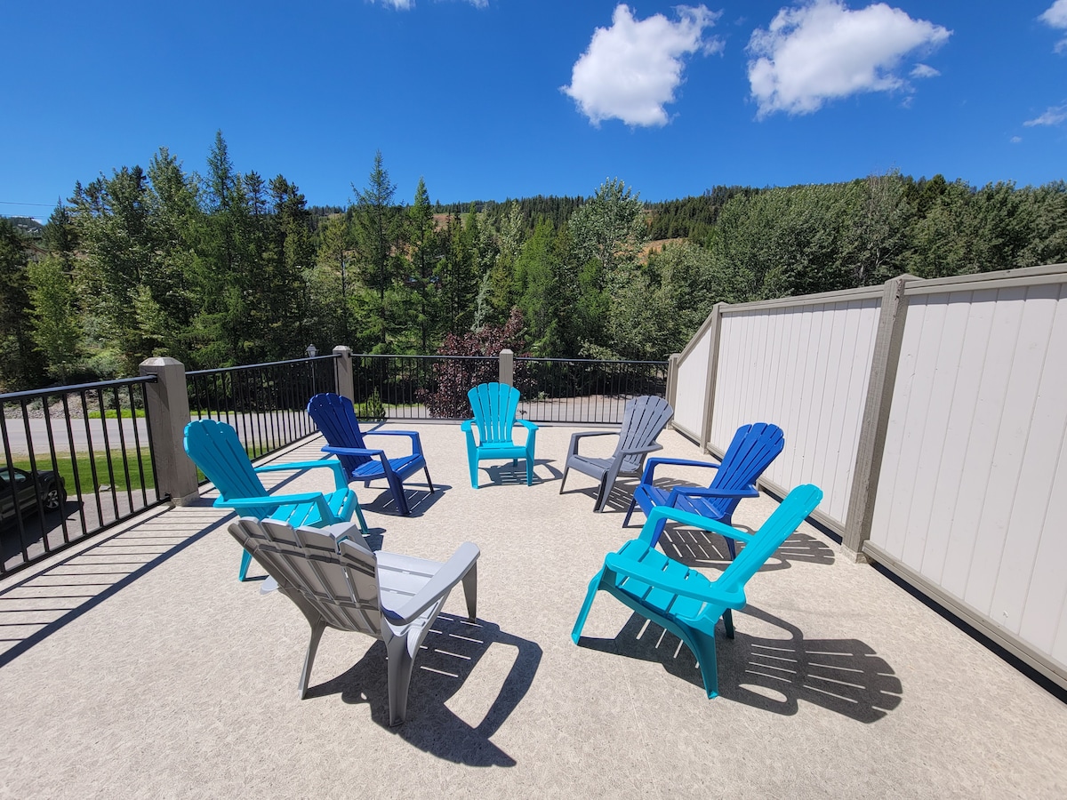 A spacious outdoor deck features six colorful lounge chairs in blue and gray, arranged for relaxation. Lush greenery from surrounding trees creates a serene backdrop, under a clear blue sky adorned with a few fluffy clouds.