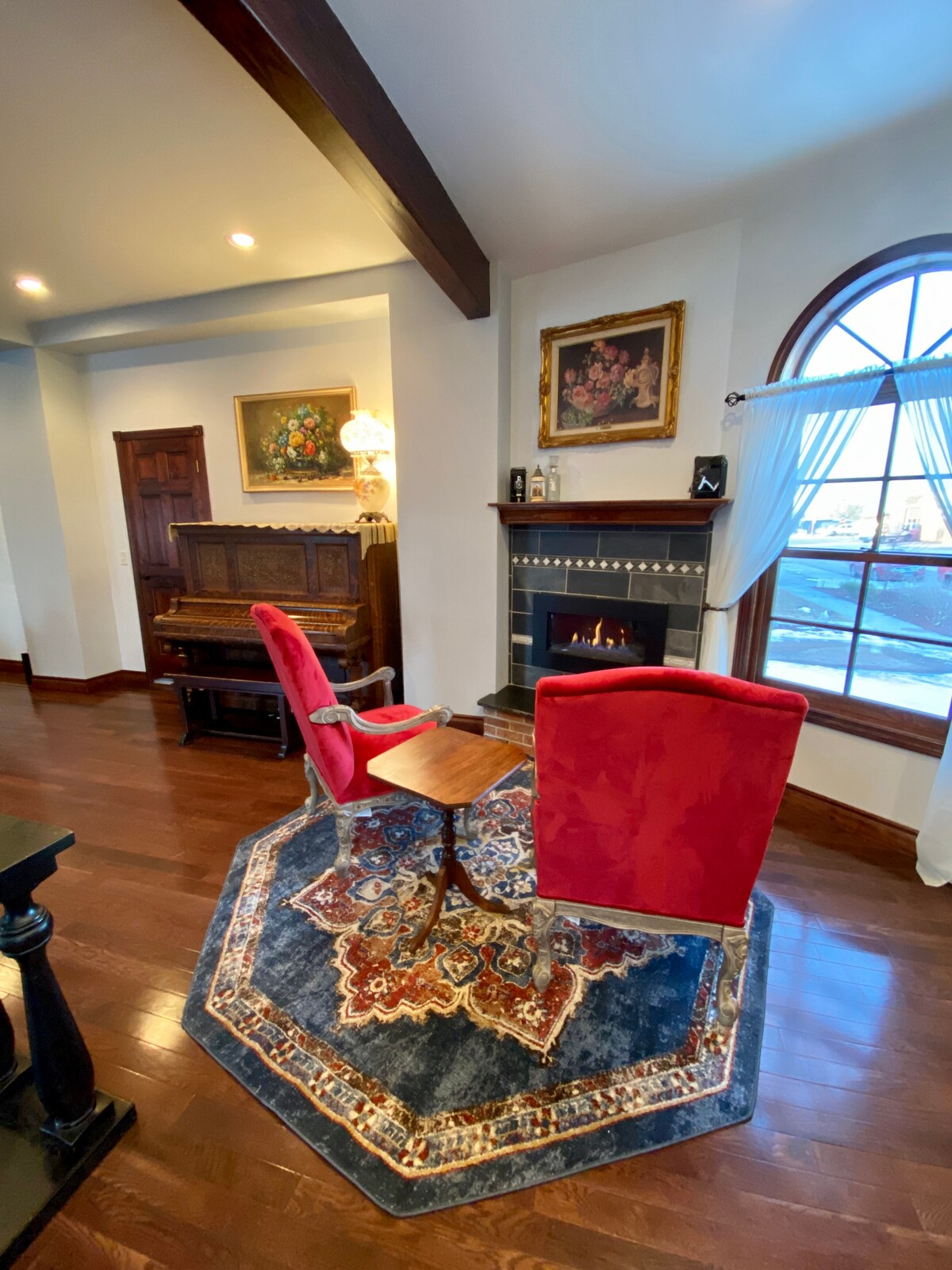 Two red armchairs are positioned around a small table on a colorful rug, creating a cozy seating area. A fireplace is set into the wall, with a painting above it and a vintage piano nearby, framed by a large arched window allowing natural light.