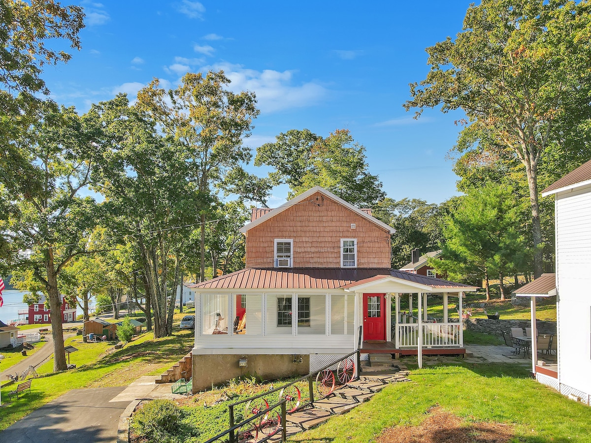 The exterior of a two-story country home features a red front door and a welcoming porch with white railings. Surrounding trees provide shade, while a path leads from the driveway to the entrance. A glimpse of the lake is visible in the background.