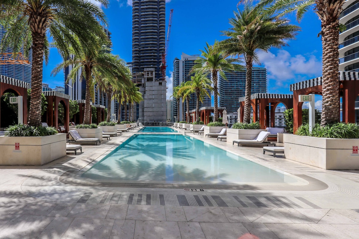 A spacious outdoor pool area is surrounded by palm trees and modern architecture. Comfortable lounge chairs are placed along the perimeter, providing ideal relaxation spots. The pool invites guests with its clear blue water beneath a bright sky, framed by sophisticated cabanas and greenery.