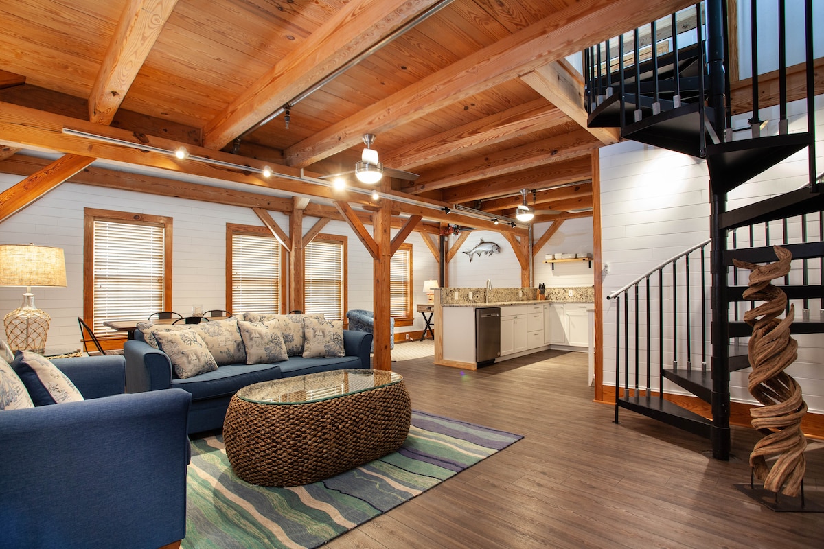 A spacious living area is highlighted by a comfortable blue sofa and a glass-top coffee table, resting on a soft area rug. Exposed timber beams and stylish light fixtures create a warm ambiance. A spiral staircase leads to the upper level, while a modern kitchen is visible in the background.