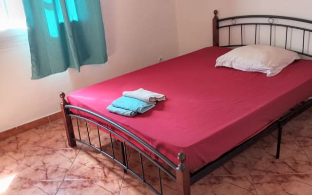 A simple bedroom features a spacious bed with a red fitted sheet and a white pillow. Towels are neatly folded on top of the bed. Soft natural light filters through a window covered with teal curtains, highlighting the tiled floor.