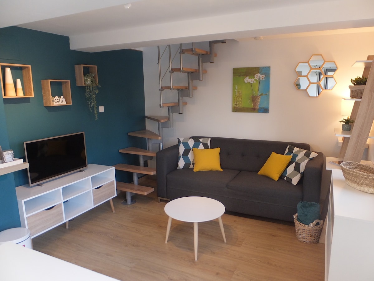 A modern living area features a comfortable gray sofa with decorative pillows in yellow and teal. A round coffee table sits in front of the sofa. Shelving and a television are arranged on one wall, while a spiral staircase leads to an upper level. Natural light highlights a colorful art piece.