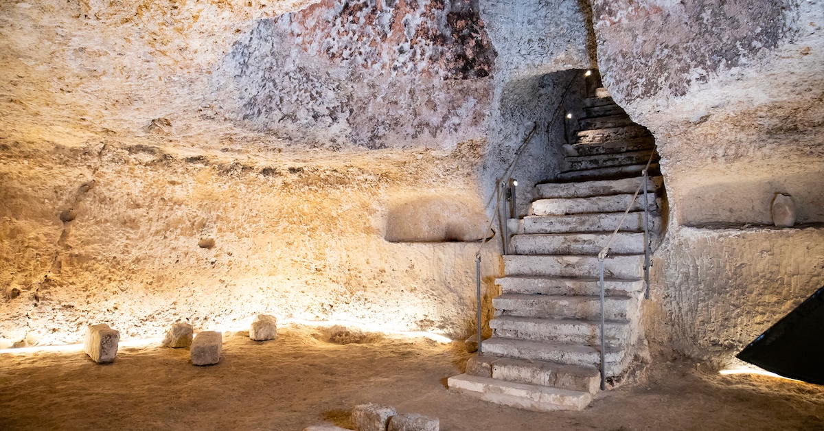 A stone staircase leads upward from a subterranean space, characterized by rough, natural walls and a subtly illuminated environment. The floor is flat and sandy, with occasional large, smooth stones placed throughout. Soft lighting highlights the texture of the rock formations.