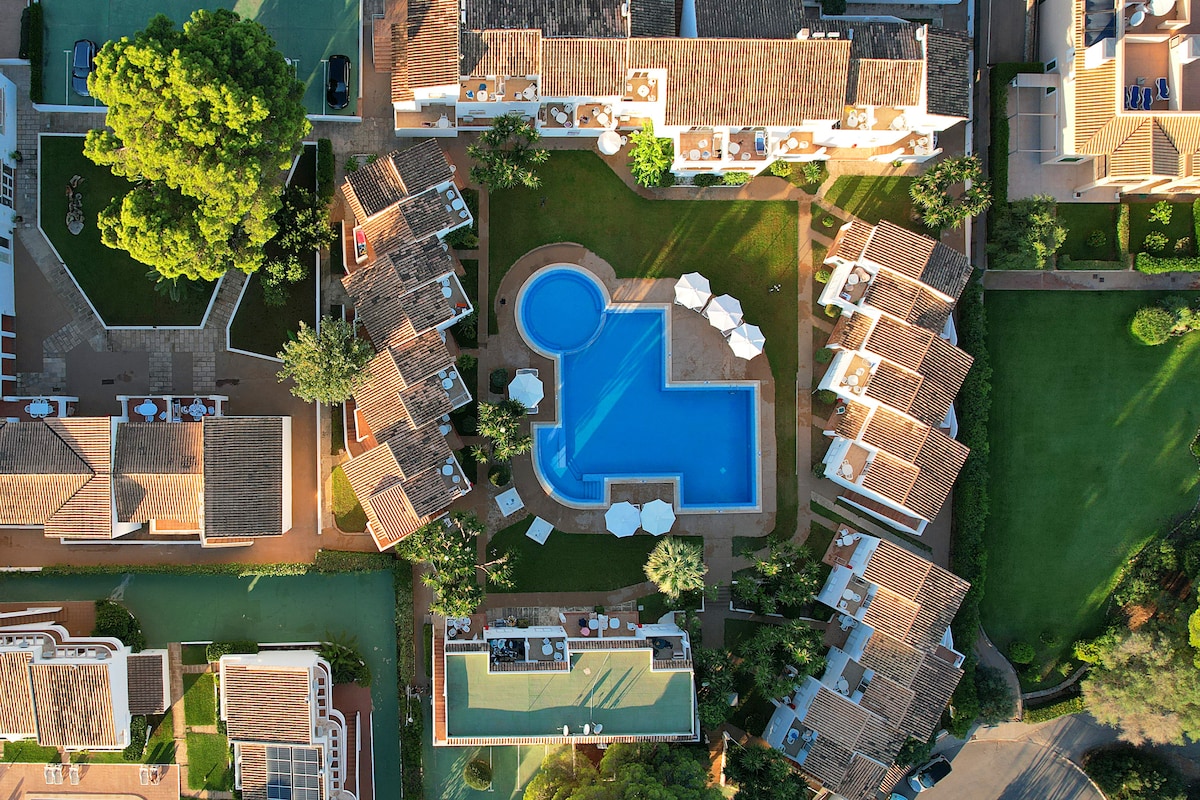 An aerial view captures the layout of the property, showcasing a central swimming pool surrounded by landscaped gardens. White lounge chairs are arranged beside the pool, while neighboring buildings exhibit terracotta roofs and green courtyards, creating a harmonious outdoor space for relaxation.